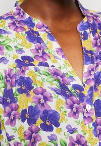 Blouse florale présentant un fond blanc, imprimé de pensées violettes et de fleurs jaunes, avec une patte de boutonnage et un col. Texture de tissu lisse.