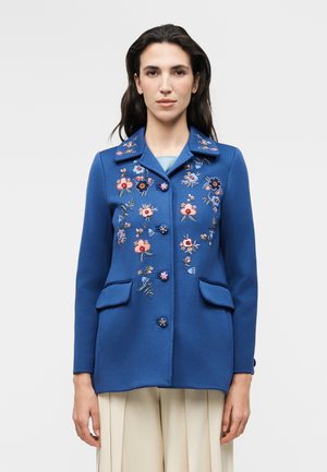Γυναίκα που φοράει μπλε σακάκι με κεντητά ρόζ και μπλε λουλουδάτα σχέδια, κουμπωμένο μπροστά, με τσέπες με καπάκι και μπεζ πλισέ παντελόνια.