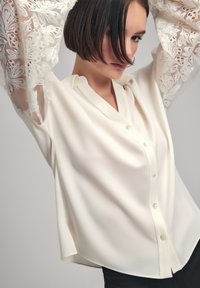 White blouse with sheer, embroidered lace sleeves, button-down front, and a relaxed fit. Smooth texture with subtle sheen.