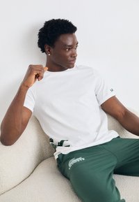Young man with short curly hair sitting on a light sofa wearing a white t-shirt and green pants with crocodile logos.