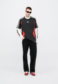 Μαύρο και κόκκινο μπλουζάκι Adidas με λευκά μακριά μανίκια από μέσα, συνδυασμένο με μαύρο παντελόνι φαρδύ και κόκκινα αθλητικά παπούτσια. Μαύρα γυαλιά ηλίου συμπληρώνουν την εμφάνιση.