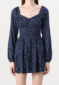 Mörkblå klänning med smockad byst, långa ärmar och vitt blommönster; har en fyrkantig halsringning och flerlagad kjol.