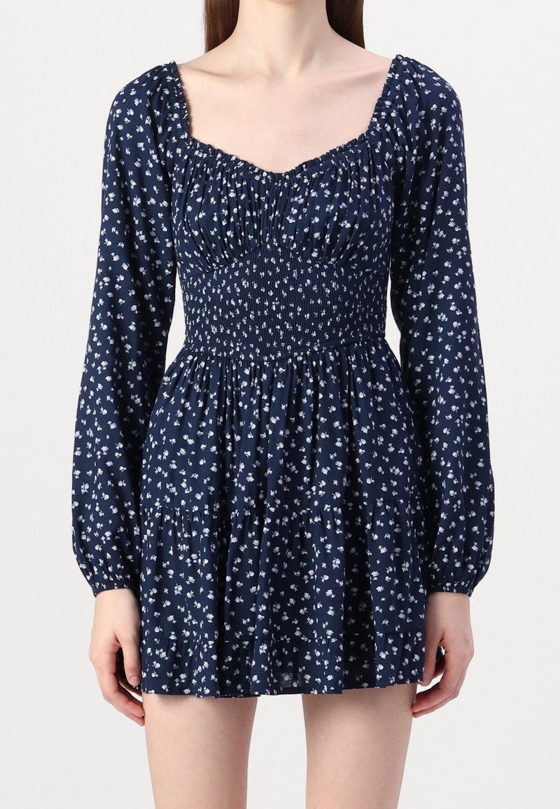 Mörkblå klänning med smockad byst, långa ärmar och vitt blommönster; har en fyrkantig halsringning och flerlagad kjol.