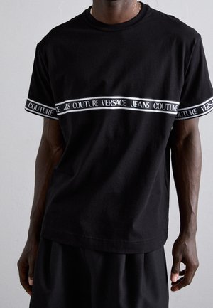 Uomo che indossa una t-shirt nera a maniche corte con strisce orizzontali bianche con il testo "Versace Jeans Couture" sul petto e sulle maniche.