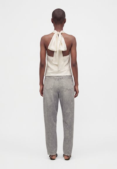 Personne aux cheveux courts portant un top crème sans manches avec un grand nœud dans le dos et un jean gris délavé taille haute, debout de dos.