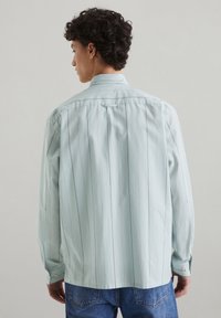 Lichtblauwe blouse met verticale witte strepen, lange mouwen, een standaard kraag en een rechte zoom. De stof lijkt glad en gestructureerd.