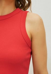Red ribbed sleeveless top featuring a high neckline and fitted design, showcasing a smooth texture and clean lines around the armhole.