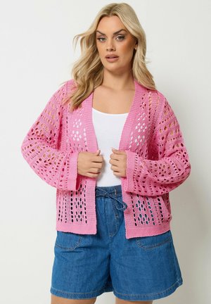 Vrouw draagt een roze opengeweven vest over een wit topje, gecombineerd met blauwe denim shorts, staand tegen een effen lichte achtergrond.
