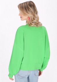 Felpa verde oversize con una texture liscia, spalle drop e orlo a coste; abbinata a jeans azzurri chiari.