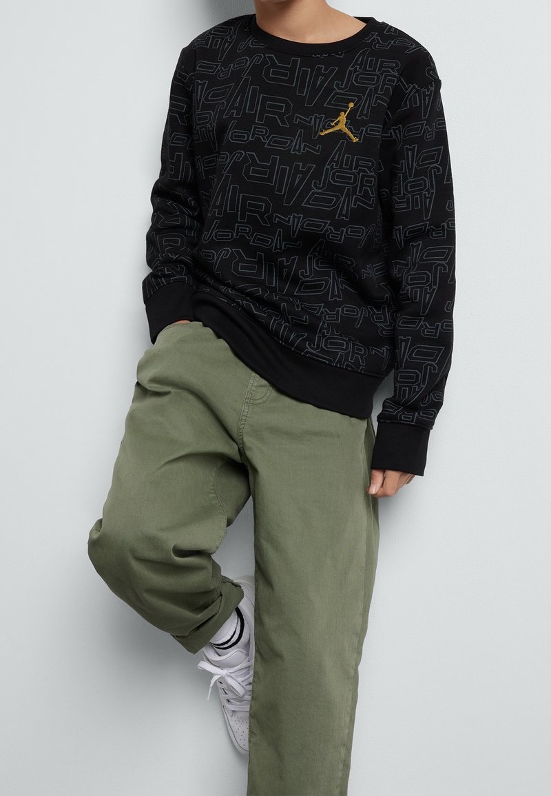 Felpa nera a maniche lunghe con motivo ripetuto del testo "Jordan" e accentuazione in oro. Pantaloni larghi verde oliva e scarpe da ginnastica bianche.
