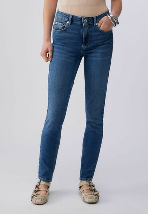 Donna che indossa jeans aderenti blu con dettagli in strass sulle tasche e scarpe Mary Jane beige con borchie, in piedi davanti a uno sfondo semplice.