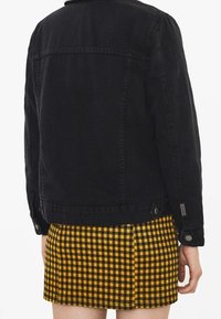 Chaqueta de mezclilla negra con un diseño estructurado, costuras visibles y dos bolsillos laterales, llevada sobre una falda corta a cuadros amarillos y negros.