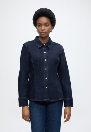 Vrouw draagt een donkerblauw spijkerjasje met zilveren knopen en bijpassende spijkerbroek, staand tegen een effen lichte achtergrond.