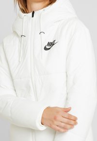 White puffer jacket featuring a zip front, adjustable hood, and black Nike logo. The material is smooth with a quilted texture.