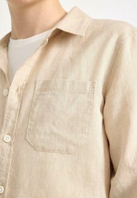 Camicia beige in misto lino con colletto a punta, taschino e bottoni bianchi, caratterizzata da una superficie morbida e textured.