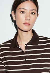 Polo marrone con strisce bianche, dotato di colletto, tre bottoni e un tessuto testurizzato. Inquadratura ravvicinata della parte superiore del torace di un modello.