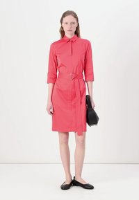 Coral shirt dress with a collared neck, three-quarter sleeves, belted waist, straight silhouette, and smooth texture, paired with a black purse.