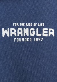 Tessuto navy con trama testurizzata e testo bianco: "PER IL VIAGGIO DELLA VITA WRANGLER FONDATA NEL 1947" in diverse dimensioni di font.