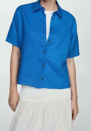 Osoba ubrana w jasnoniebieską, krótką koszulę z guzikiem na białej bluzce i białej spódnicy na jednolitym tle.