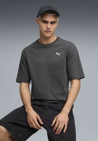 Camiseta de algodón gris oscuro de manga corta, con un logo blanco de Puma en el lado izquierdo del pecho. Combinada con pantalones cortos de mezclilla negros, en una pose sentada.