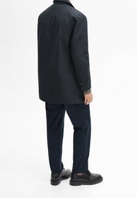 Mustat ulkoilutakit, joissa on kaulus, nappidetalit ja löysä istuvuus, yhdistettyinä tummansinisiin housuihin ja mustiin kenkiin, nähtynä takaa.