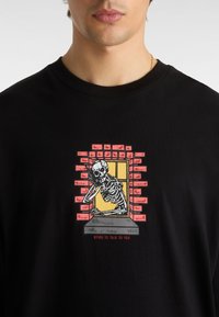 Schwarzes Baumwoll-T-Shirt mit einem Skelettmotiv in einem Fenster, das von roten Ziegeln umrahmt ist, und dem Text "DYING TO TALK TO YOU" darunter.