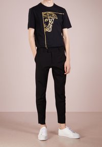 Camiseta de algodón negra con diseño gráfico dorado de una cara; combinada con pantalones negros a medida y zapatillas bajas blancas. Textura suave.