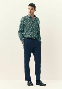 Mand iført en grøn blomstret skjorte med lange ærmer, navy blå bukser, sort bælte og sorte loafers, står med hænderne i lommerne foran en ensfarvet baggrund.