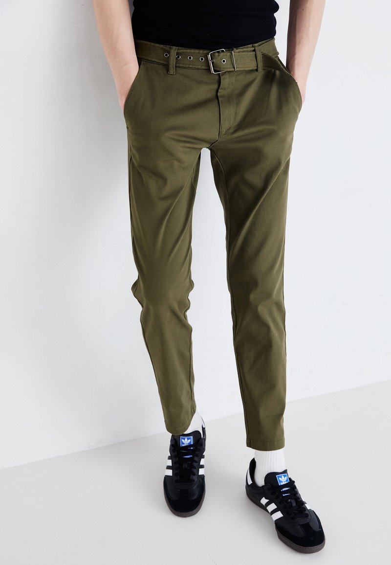 Olivenfargede chinos laget av bomull, med belte med sølvspenne, sidelommer og avsmalnende ben, kombinert med svarte sneakers.
