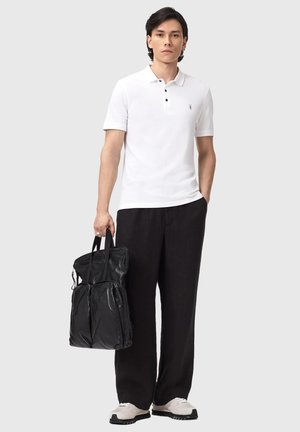 Mann trägt weißes Polohemd, schwarze weit geschnittene Hose, weiße Turnschuhe, hält schwarze Ledertasche mit zwei Vorderfächern, steht vor einfarbigem Hintergrund.