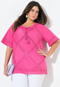 Blusa rosa con maniche corte, caratterizzata da dettagli in pizzo e accenti di nappine. Indossata con pantaloni bianchi e una borsa lilla chiaro.