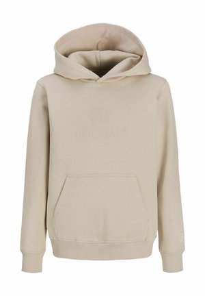 Garçon portant un sweat à capuche beige, une chemise à carreaux, un jean large foncé, des baskets beiges et une casquette de baseball noire, regardant vers sa droite sur fond blanc.
