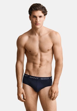 Jeune homme à la silhouette musclée portant un slip bleu marine de la marque GANT, debout devant un fond clair uni.