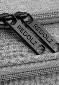 Tela gris texturizada con cremalleras negras que presentan la marca "REDOLZ". Primer plano que muestra los dientes de la cremallera y los detalles de costura.