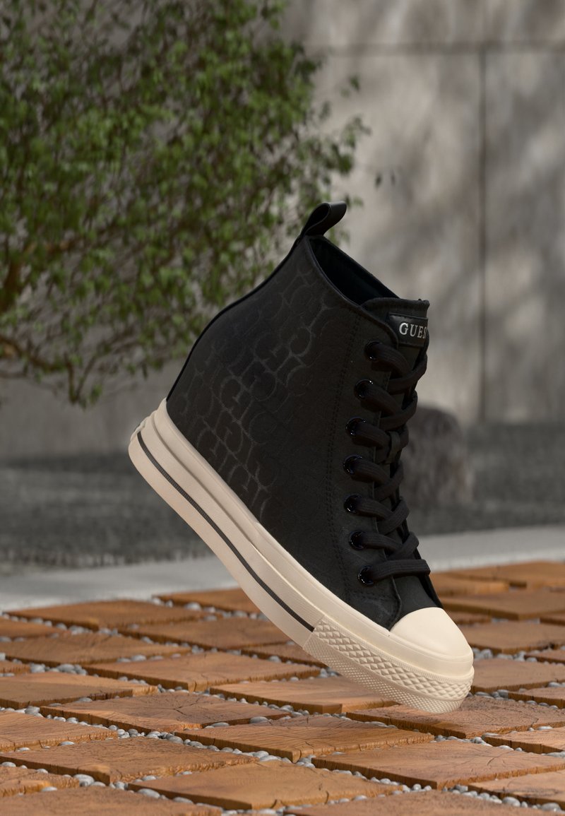 Μαύρο sneaker με ψηλή σόλα, λευκή σόλα και μύτη, με διακριτικό μοτίβο και λογότυπο "GUESS" στη γλώσσα, τοποθετημένο όρθιο σε ξύλινη επιφάνεια.