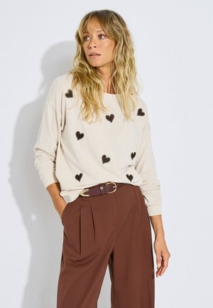 Mujer rubia con cabello ondulado lleva un suéter crema con corazones negros y pantalones marrones de cintura alta con un cinturón oscuro, mirando hacia un lado.