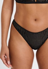 Slip bikini nero in tessuto liscio con dettagli a righe verticali, caratterizzato da un design a vita bassa e copertura minima.