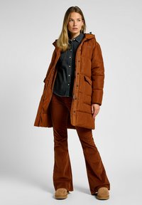 Brun polstret jakke med hætte, der har lynlås og knaplukning, kombineret med en mørk denimskjorte og brune flared bukser i kordfløjl.