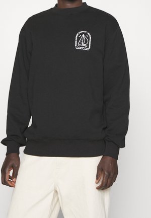 Czarna bluza z białym logo jachtu i napisem "Manga Clothing Company - Helmsman Harbour" na piersi, noszona z jasnymi spodniami.