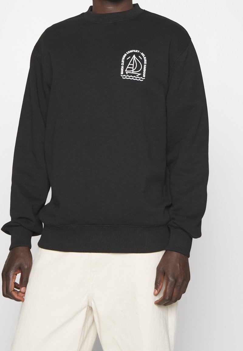 Sweat noir avec logo de voilier blanc et texte « Manga Clothing Company - Helmsman Harbour » sur la poitrine, porté avec un pantalon clair.