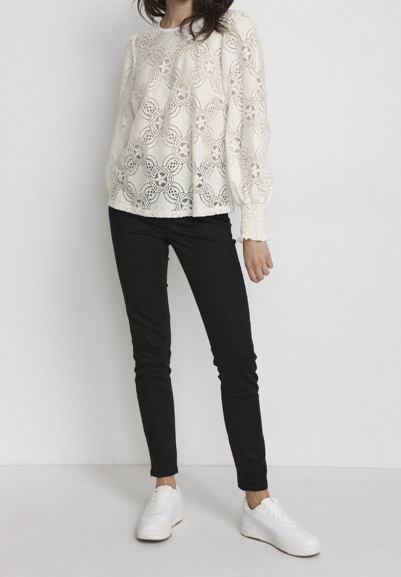 Blouse en dentelle à manches longues crème, ornée de motifs floraux et avec des manches bouffantes, assortie à un jean noir ajusté et des baskets blanches.