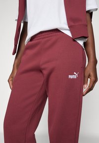 Burgundy træningsbukser lavet af tekstureret stof, med elastisk talje og hvid Puma-logo på venstre lår.
