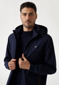 Giacca a zip blu navy realizzata in tessuto liscio, con cappuccio, polsini elasticizzati e un logo sul petto. Design minimalista.