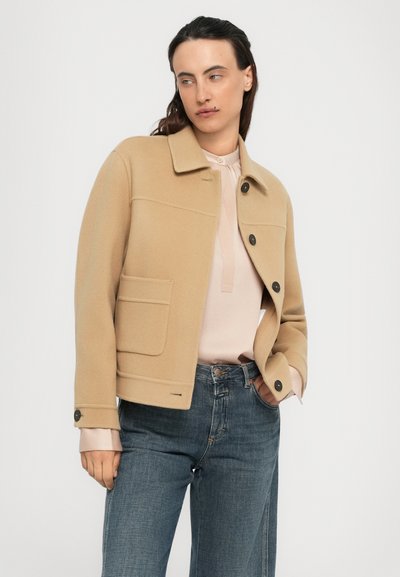 Person in einer beigen Knopfjacket über einer blassroten Bluse und blauen Jeans, steht mit einer Hand in der Tasche vor einem schlichten Hintergrund.