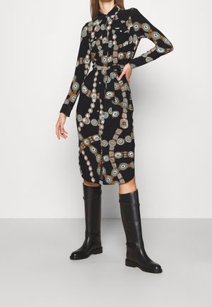 Woman wearing a black long-sleeve belted dress with a colorful chain pattern and black knee-high boots, standing against a white wall.