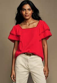 Top in cotone rosso con maniche corte arricciate e scollatura quadrata; presenta un tessuto texturizzato e delicati dettagli in pizzo. Abbinato a pantaloni crema.