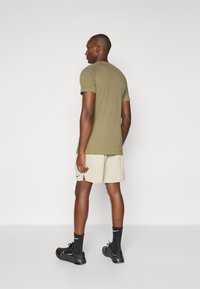 Olivgrünes, eng anliegendes T-Shirt kombiniert mit hellbeigen Shorts, versehen mit einem Nike-Logo, schwarzen Socken und schwarzen Sportschuhen. Einfaches Design.
