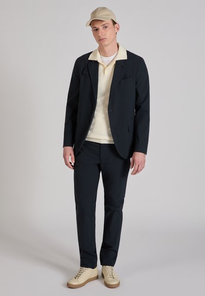 Man die een beige pet, wit shirt, crèmekleurige trui, zwarte blazer, zwarte broek en crèmekleurige sneakers draagt, staand tegen een effen grijze achtergrond.