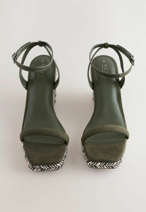 Olivgröna högklackade sandaler med fyrkantiga tår, ankelsnoddar och vävd svart-vit detalj på sulan. Material i mocka.