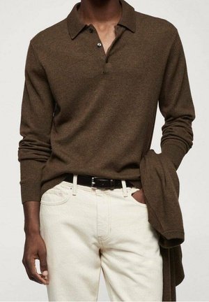 Mand iført brun langærmet poloshirt stoppet ned i off-white bukser med sort bælte, der holder en matchende brun sweater i venstre hånd.